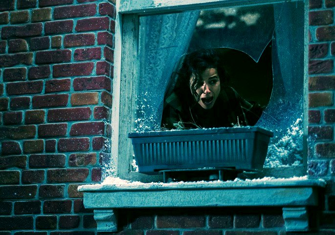 Una mujer gritando en una ventana de una casa con vidrios rotos.