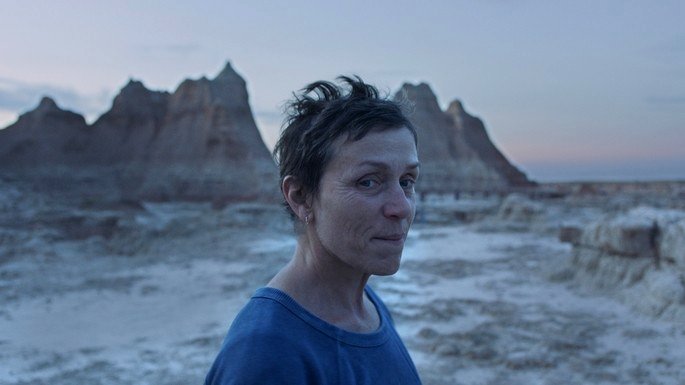 Una mujer de pelo corto y blusa azul, con un desierto y montañas de fondo