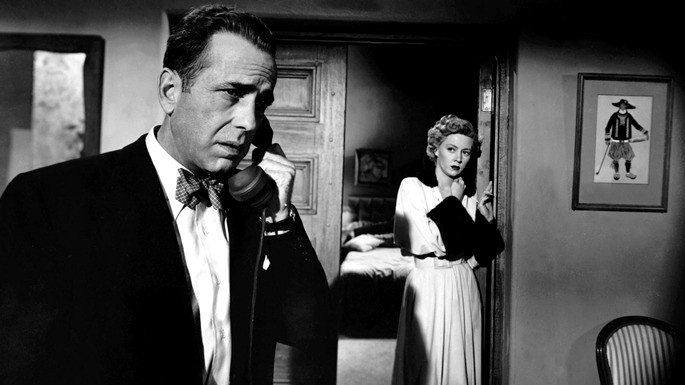 Imagen en blanco y negro con un hombre hablando por teléfono y, de fondo, una mujer escuchando detrás de la puerta
